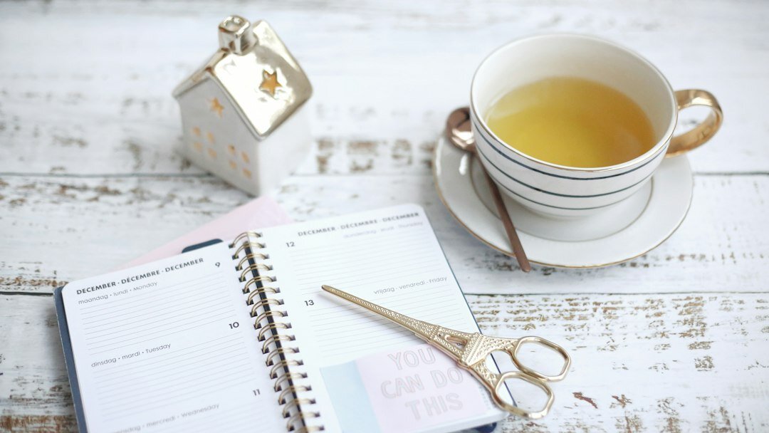 Flat lay photo of a desk with a calendar planner, notebook, pen and a cup of green tea